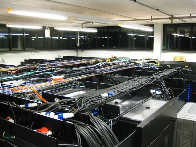 Rows of server racks in a computer room, their tops covered in thick black power cables and orange and yellow fibre leads.